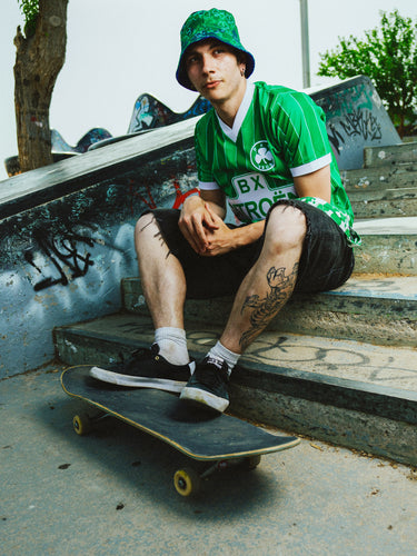 Person in Panathinaikos shirt and hat sitting on a skateboard with graffiti in the background