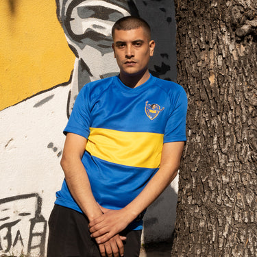 Model wearing the Maradona x COPA Boca 1981 - 82 Retro Football Shirt, posing against a mural background.