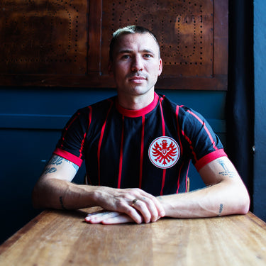 Eintracht Frankfurt 1972 - 73 Retro Football Shirt Black & Red Lifestyle worn by a model sitting in a dimly lit indoor setting.