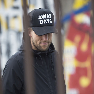 Man wearing a black "AWAY DAYS" COPA trucker cap standing near a graffiti wall