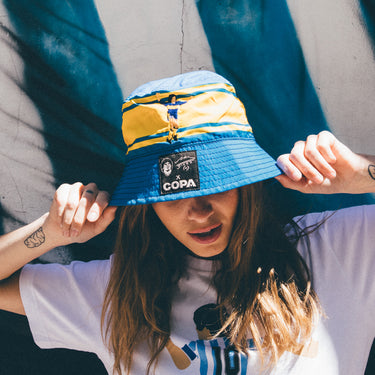 Model wearing a blue and yellow COPA bucket hat with embroidered Maradona design, adjusting the brim outdoors