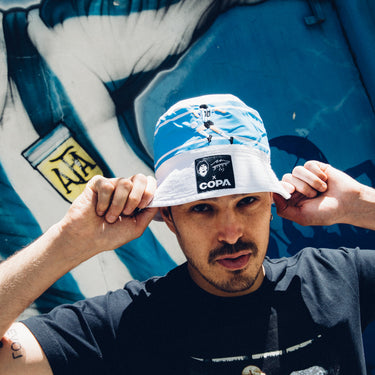 Model wearing a white and blue COPA bucket hat with embroidered Maradona design, adjusting the brim outdoors