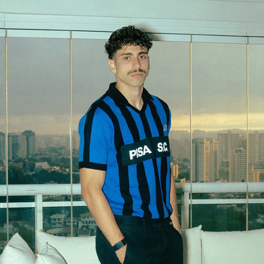 Man wearing a blue and black Pisa S.C. retro football shirt on a balcony overlooking a cityscape.
