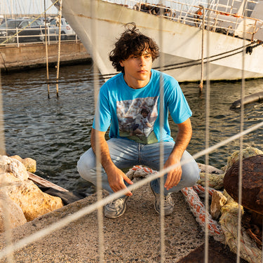 Lifestyle shot of a model wearing the Maradona x COPA Napoli Away T-Shirt, sitting near a harbor.