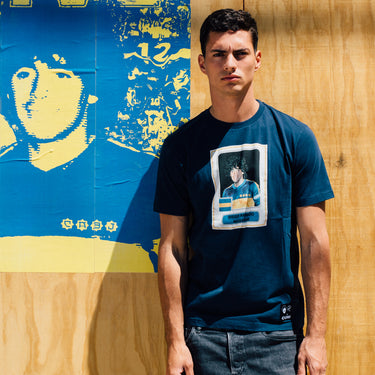 Model wearing the Maradona x COPA Boca Football Sticker T-Shirt, standing against a blue and yellow background with Maradona artwork.
