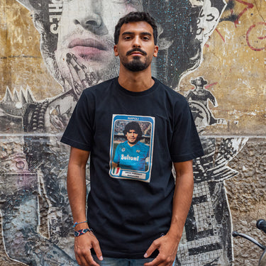 Model wearing the Maradona x COPA Napoli Football Sticker T-Shirt, standing in front of a mural featuring Diego Maradona.