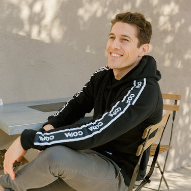 Model sitting outdoors wearing the black COPA hoodie with visible sleeve branding.
